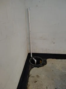 A basement corner with a sump pump pit, PVC pipe, and waterproofing system along the walls and floor. The concrete floor has some discoloration.