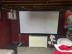 Basement home theater setup with a large projector screen on a maroon wall. Below the screen is a cabinet. To the right, there's a Christmas tree partially visible.